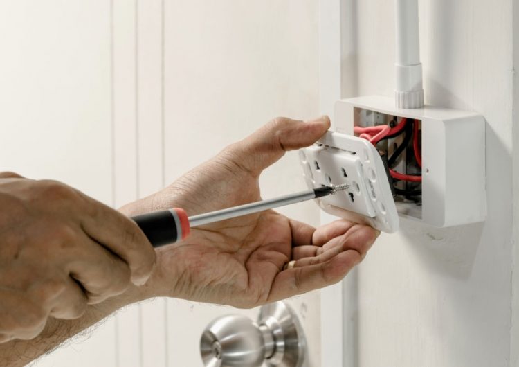 The electrician is using a screwdriver to attach the power cord to the wall outlet.